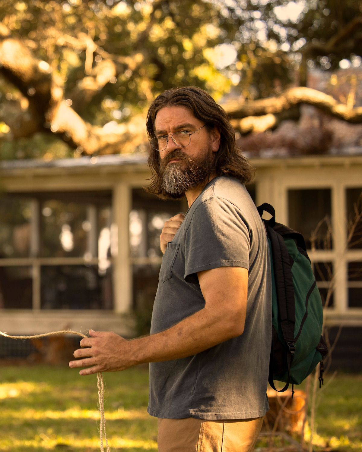 Charles Halford as John “Big John” Routledge, with long hair wearing glasses and a t-shirt stands outdoors with a backpack, in front of a house surrounded by trees and evening sunlight.