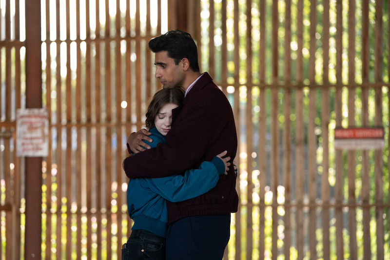A man and a child share a comforting hug in front of a tall metal fence outdoors, with sunlight filtering through the vertical bars in the background.