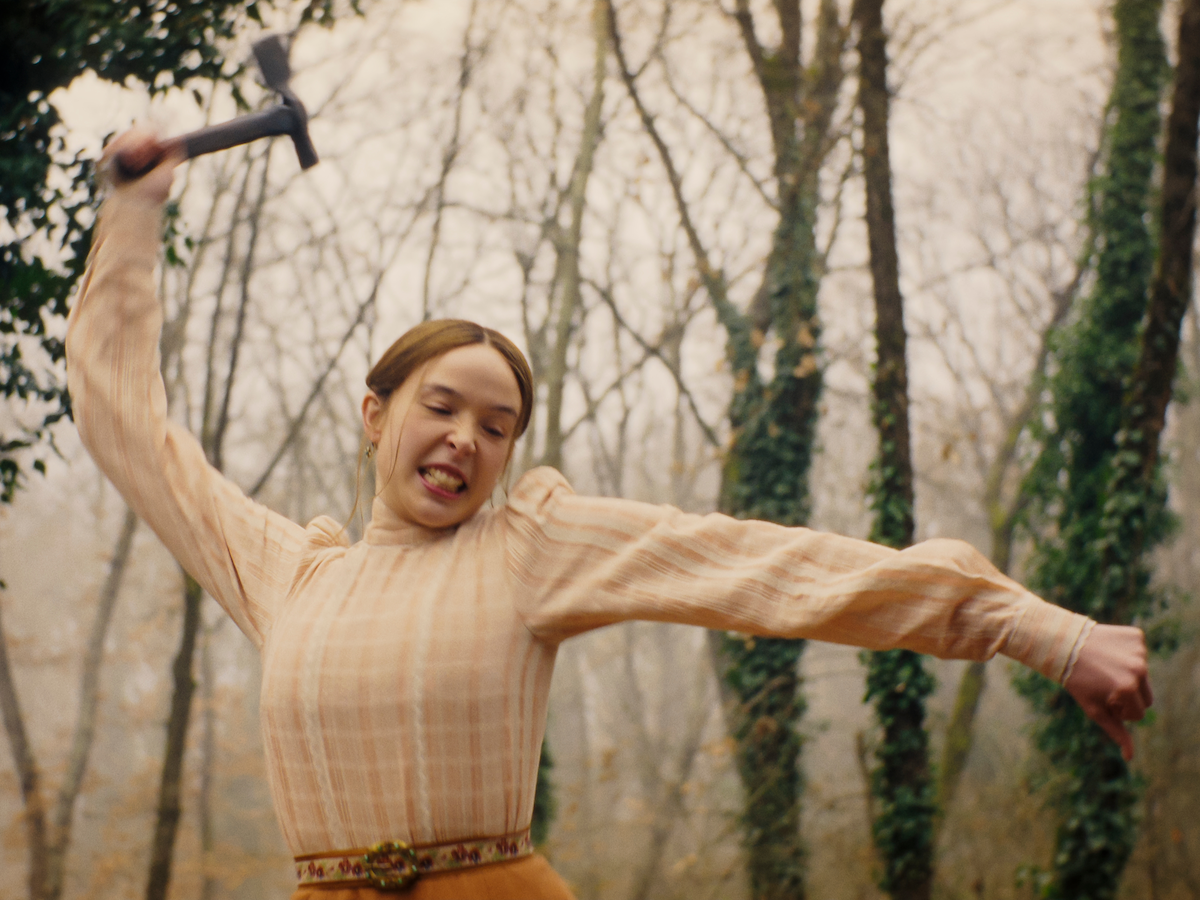 Young woman in a vintage dress swinging a hammer with intense expression in a forest, surrounded by bare trees covered in ivy on a misty day.