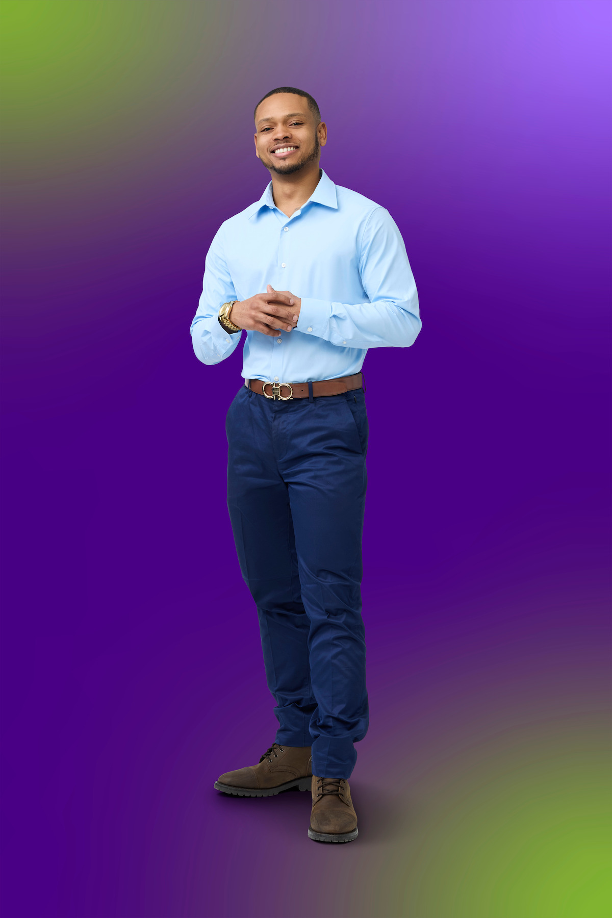 Smiling man in a light blue shirt and navy pants stands against a gradient purple background, hands clasped, in a studio setting with soft lighting.