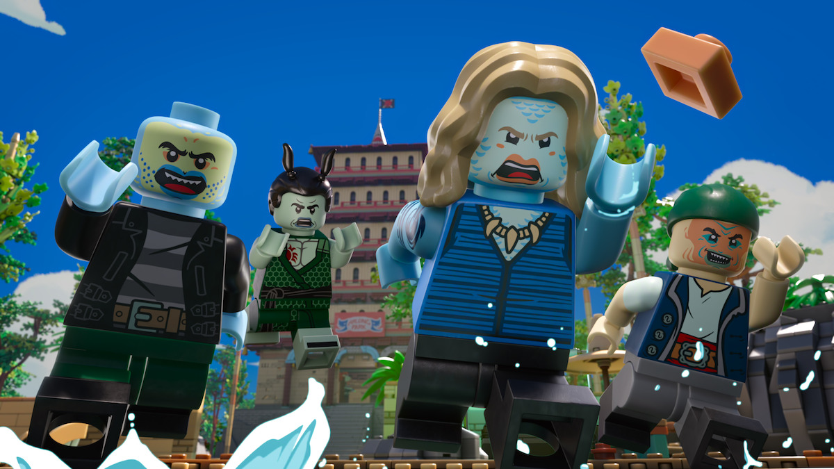 Four LEGO pirate and villain minifigures run forward outdoors, with water splashing in front of them, tropical palm trees, and a large building in the background under a blue sky. One figure throws a LEGO brick.