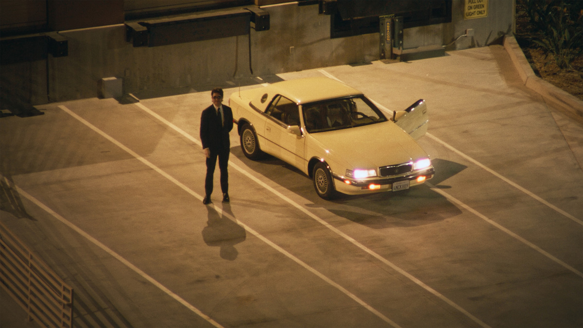 John Mulaney stands next to a white car in an empty parking lot.