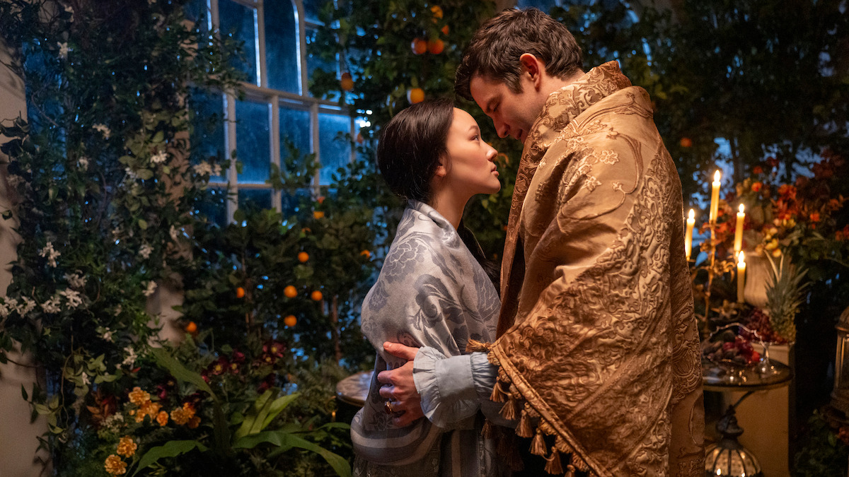 A man and woman in ornate robes share an intimate moment in a lush, candlelit indoor garden, surrounded by plants, flowers, and fruits near a large window at night.
