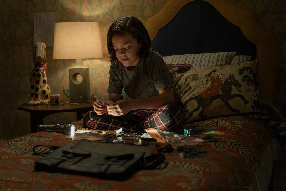 Child sitting cross-legged on a bed in a dimly lit bedroom, focused on crafting or assembling items spread out in front of them, with a lamp and decorative objects on the bedside table and patterned bedding and pillows.