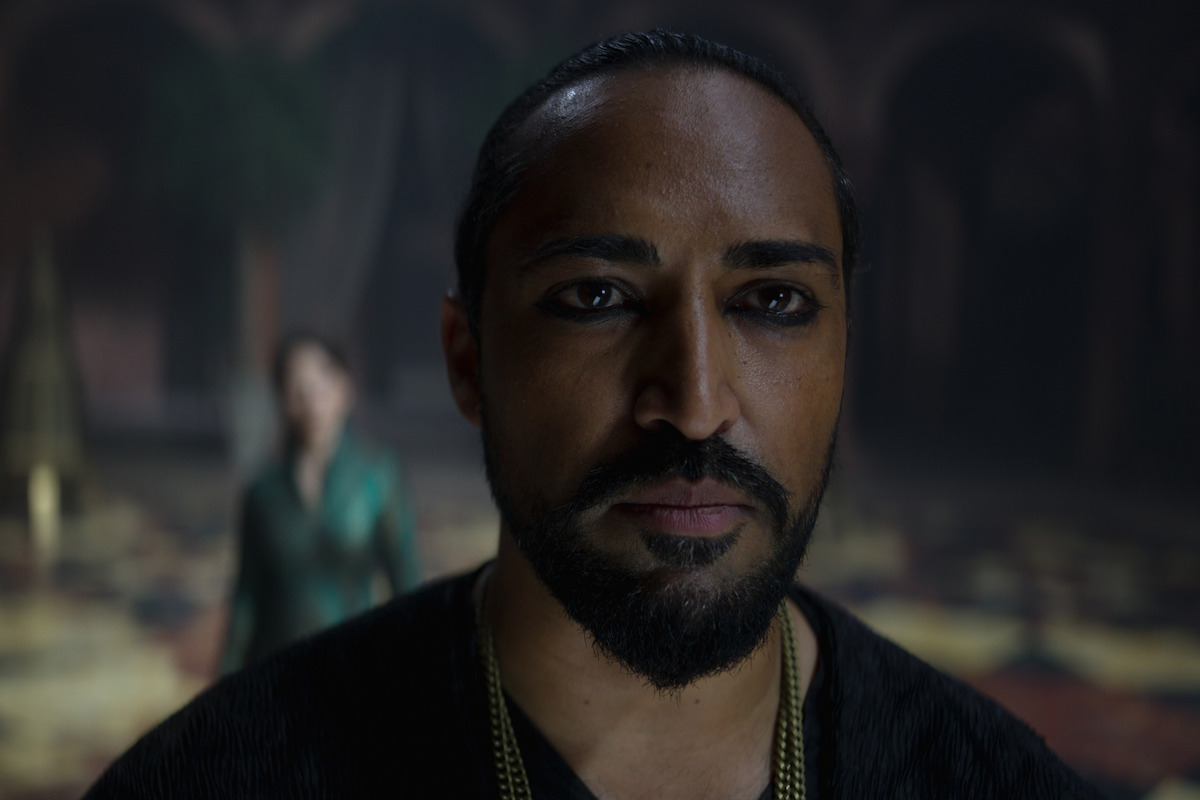 A serious man with dark eyeliner and a beard stands in sharp focus indoors, while a woman in the background is blurred. The lighting is moody, creating a tense and dramatic atmosphere.