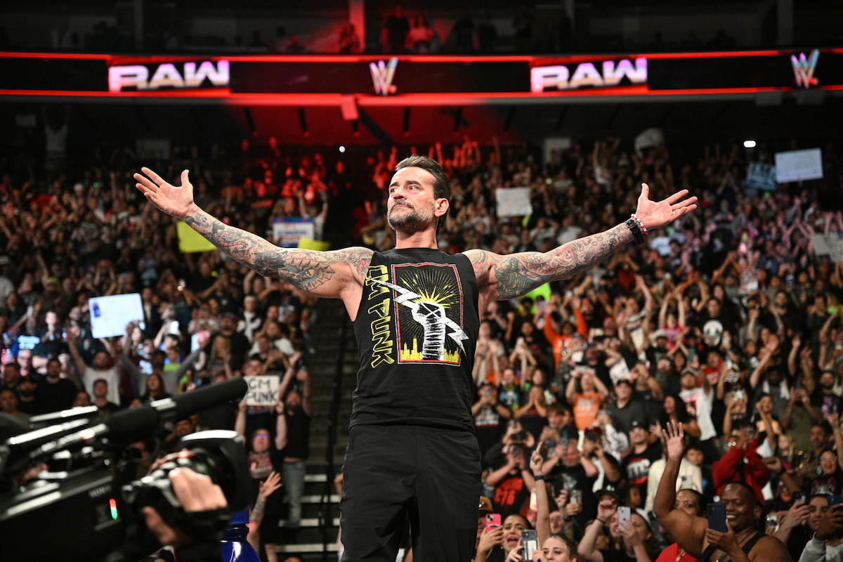 CM Punk with arms outstretched stands confidently in a packed arena during a WWE Raw event, with a large, cheering audience and bright lights in the background.