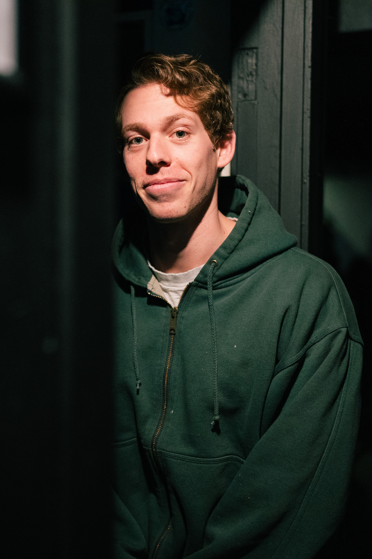 Young man with light brown hair wearing a green hoodie stands indoors, lit by dramatic side lighting, with a neutral expression and a dark background with vertical wooden panels.