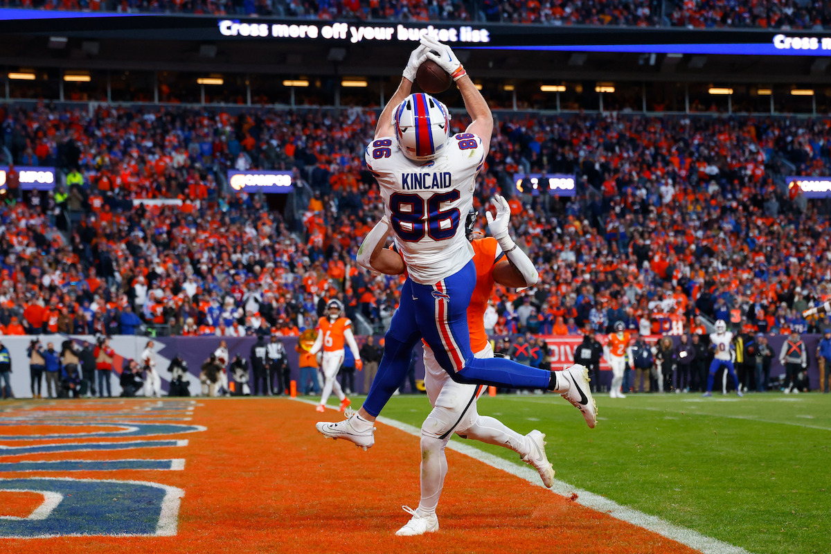 Football player in white uniform jumps to catch a pass in the end zone during a game, with a defender closely guarding him. Stadium is filled with fans and bright lights, creating an intense sports atmosphere.