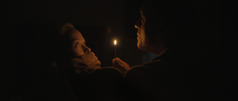 In a dark setting, a man holds a lit match close to his face while covering a woman's mouth, both illuminated by the small flame.