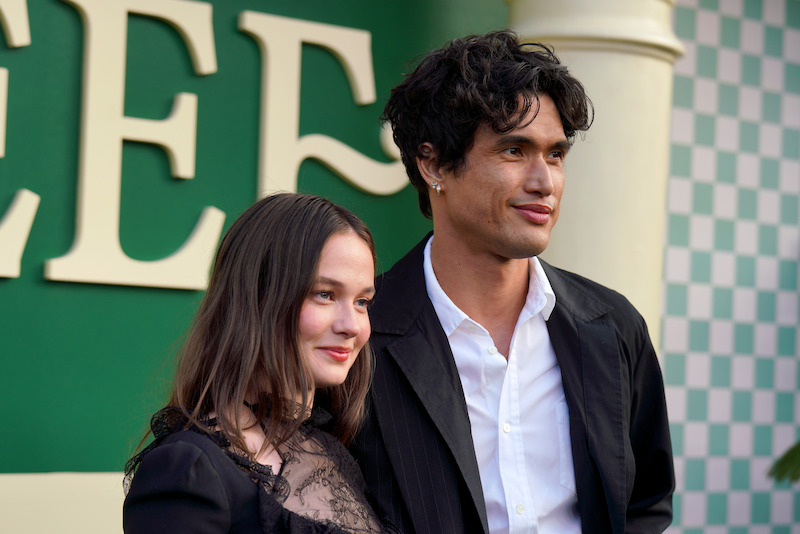 Two people dressed formally pose together, smiling, in front of a green and white backdrop with large letters and checkered accents, suggesting a public event or premiere setting.