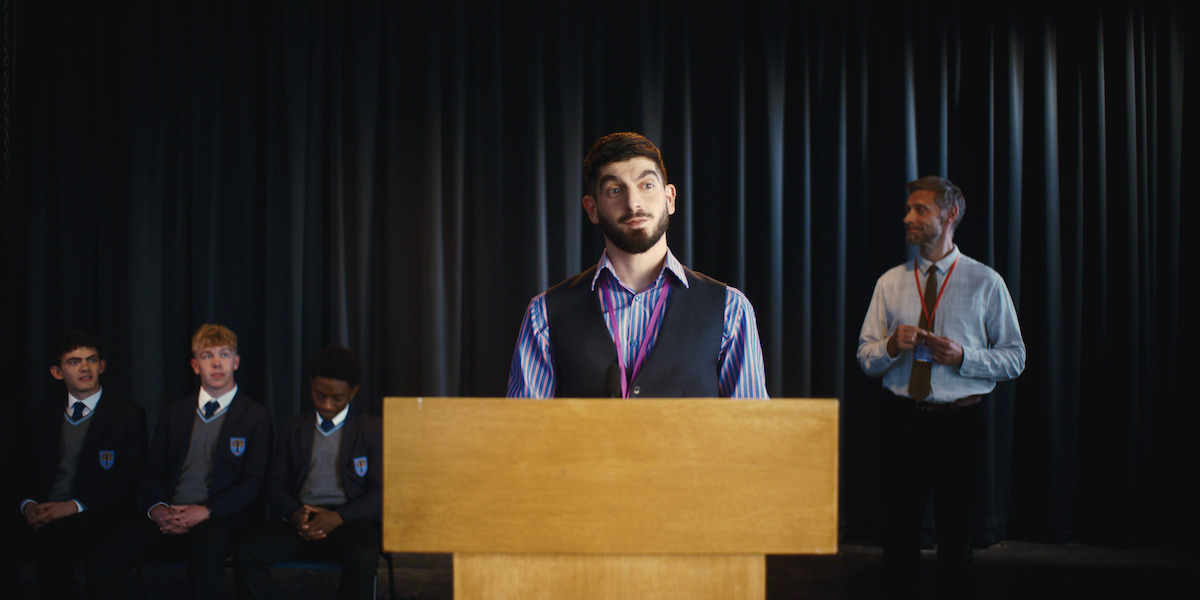 A young man stands at a podium on stage, speaking or preparing to speak, with four seated students in school uniforms and an adult standing in the background, all in front of dark curtains, suggesting a school assembly or event.