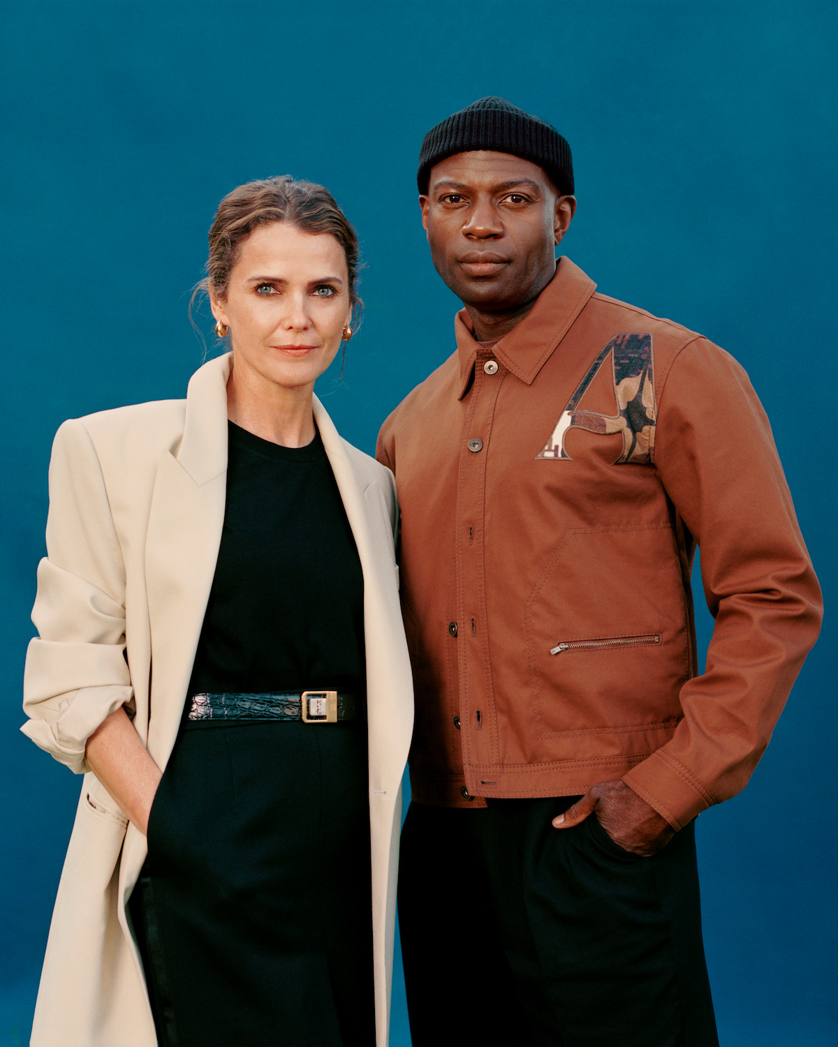 Keri Russell, wearing a white coat and the black top and pants, standing next to David Gyasi, who is wearing a black hat and the brown jacket, in front of a blue background.