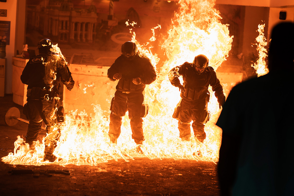 Three riot police in protective gear stand in flames indoors, fire surrounding them in what appears to be a chaotic protest or emergency situation, with a silhouette of a bystander in the foreground.