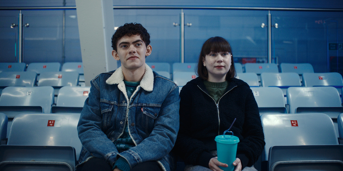 Two people sit in empty stadium seats; the young man wears a denim jacket and the young woman holds a large drink cup. The environment is bright with blue seating and glass panels in the background, suggesting an indoor setting.