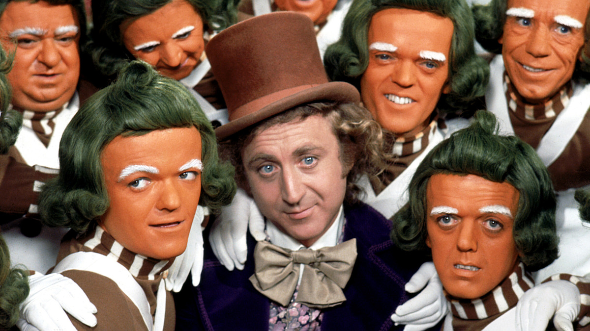 Man in a brown top hat surrounded by people with orange faces, green hair, and striped outfits, posing together closely in a whimsical, colorful setting reminiscent of a fantasy or candy factory environment.