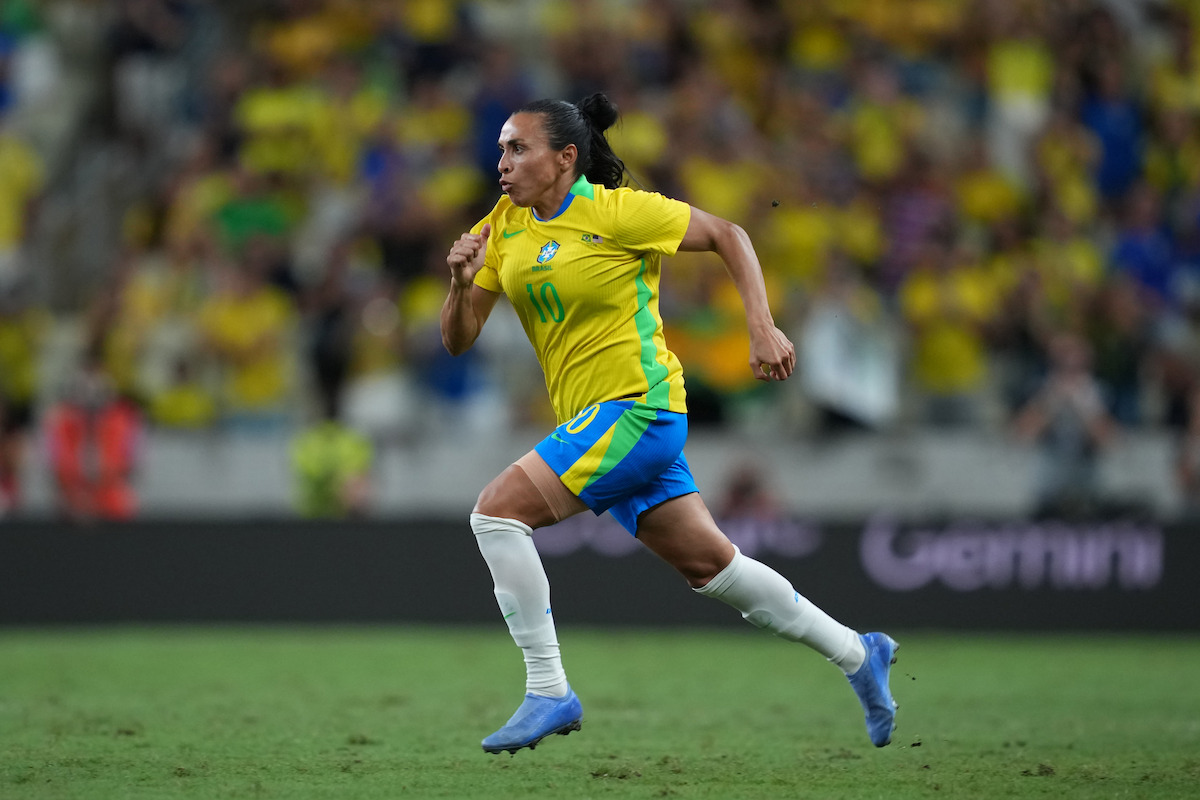 A female soccer player in a yellow and blue uniform runs on a grass field during a match in a crowded stadium, focused on the game, with fans blurred in the background.