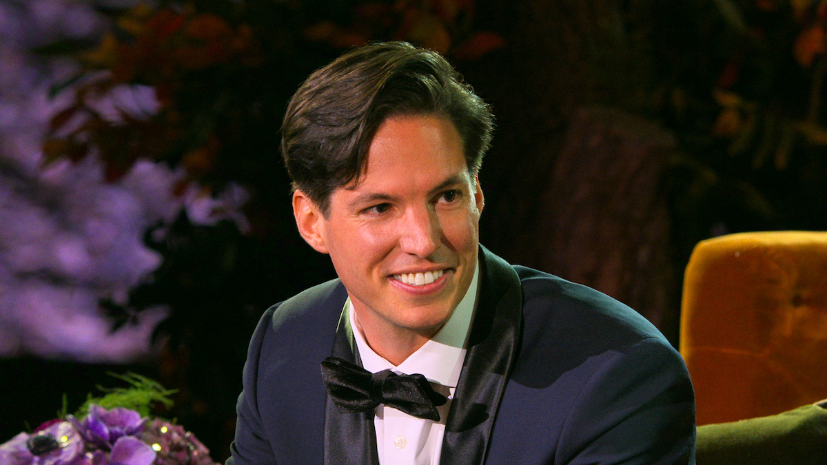 A man in a formal tuxedo sits indoors on a golden chair, smiling warmly. The background features soft, colorful lighting and decorative flowers, giving the scene a festive and elegant atmosphere.