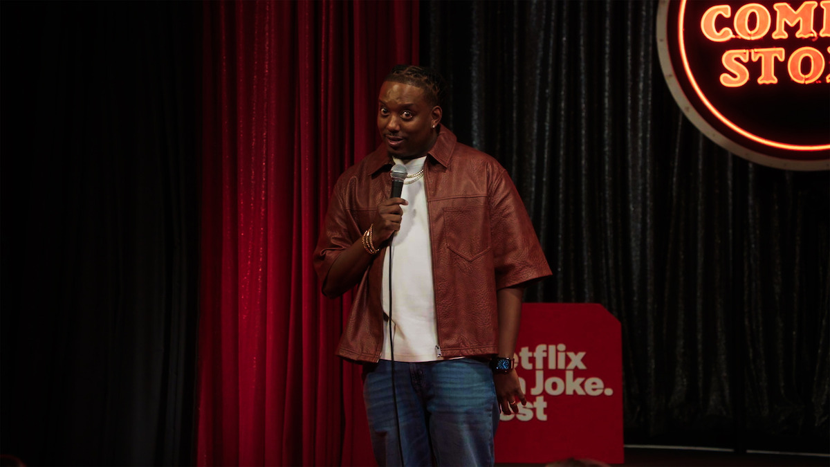 Comedian performing stand-up on stage, holding a microphone. Red and black curtains, a neon "Comedy Store" sign, and a "Netflix is a Joke Fest" sign in the background. Indoor comedy club environment.