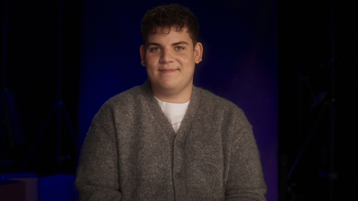 A young person with short curly hair wearing a gray sweater sits and smiles in a dimly lit studio featuring a dark blue and purple background.