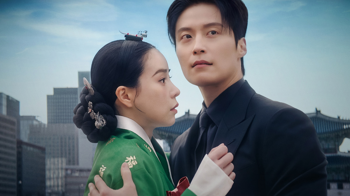 A woman in traditional Korean hanbok and a man in a modern black suit embrace with concern, standing outdoors against a backdrop of city skyscrapers and historical palace architecture under a blue sky.