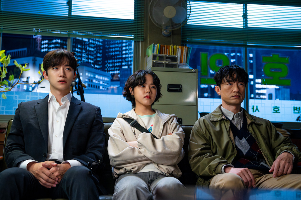 Three people sit side by side in an office with city lights visible through the blinds behind them, a man in a suit, a young woman with arms crossed, and a man in a sweater and jacket, all appearing serious.