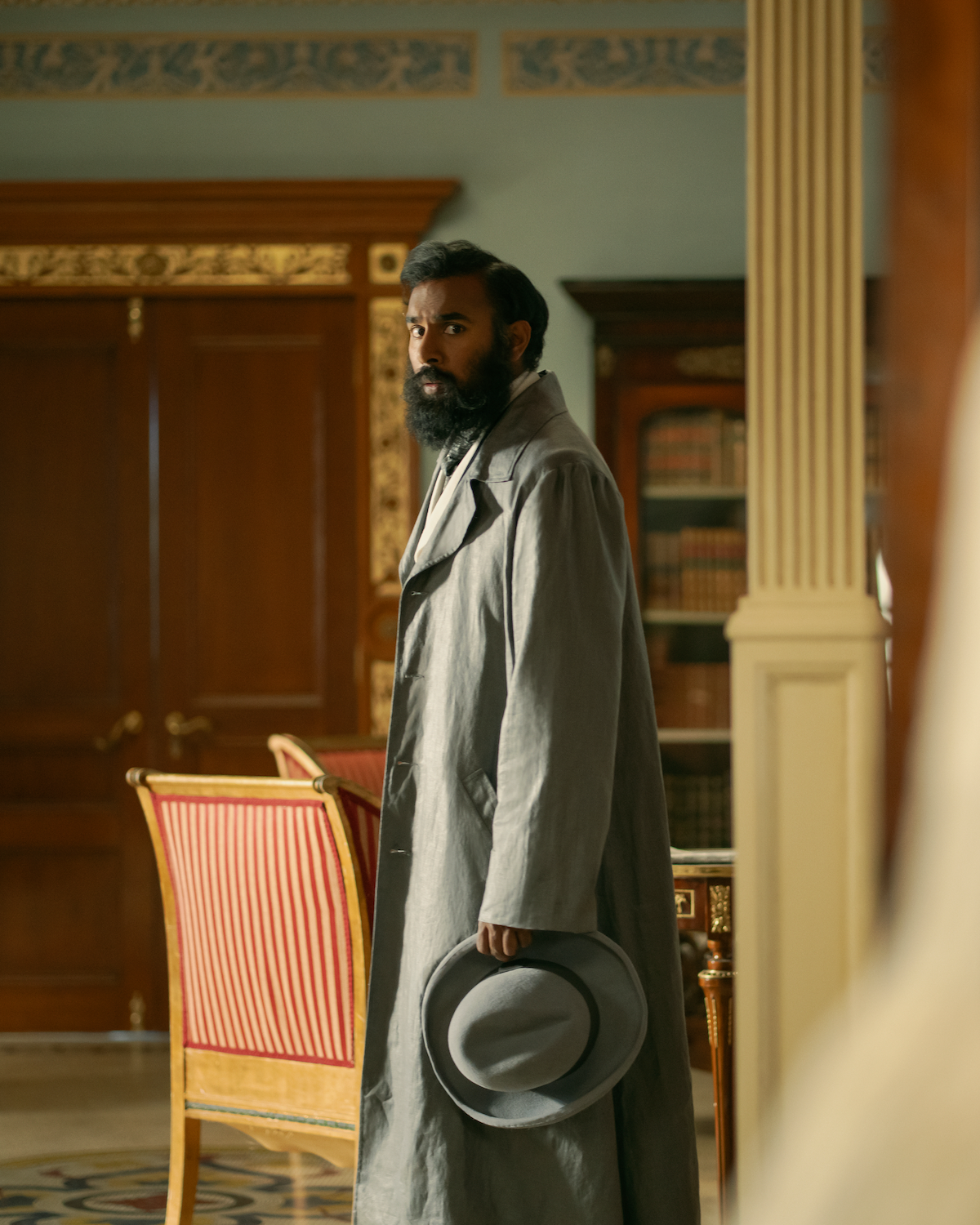 Man with a beard in a long coat holding a hat stands in an ornate room with wooden doors, gold accents, and bookshelves, looking back over his shoulder, with red upholstered chairs in the background.