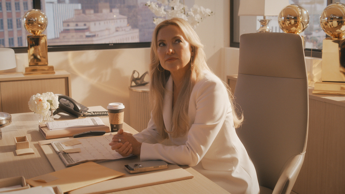 Woman with long blonde hair in a white suit sits at a modern office desk with paperwork, coffee cup, phone, and golden trophies, with cityscape visible through large windows in the background.