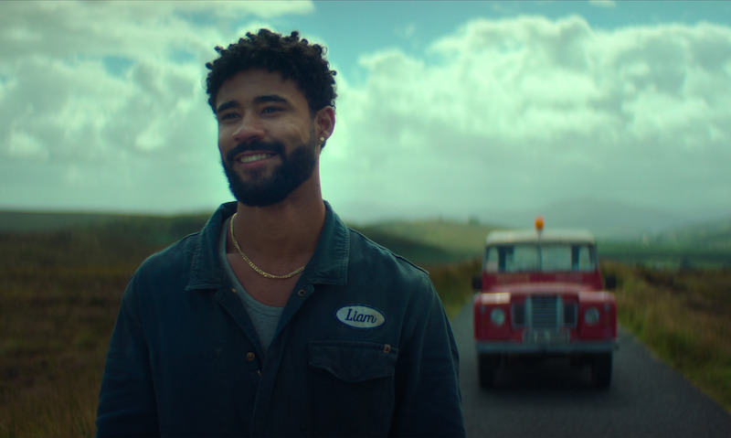 A smiling man stands on a rural road with grassy fields and mountains in the background, wearing a work shirt labeled "Liam." A vintage red truck is parked behind him. The mood is calm and uplifting.