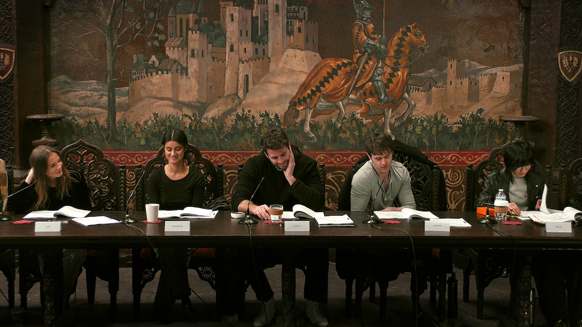 The cast of Season 4 of 'The Witcher' sits at a long table reading scripts with a tapestry hanging behind them.