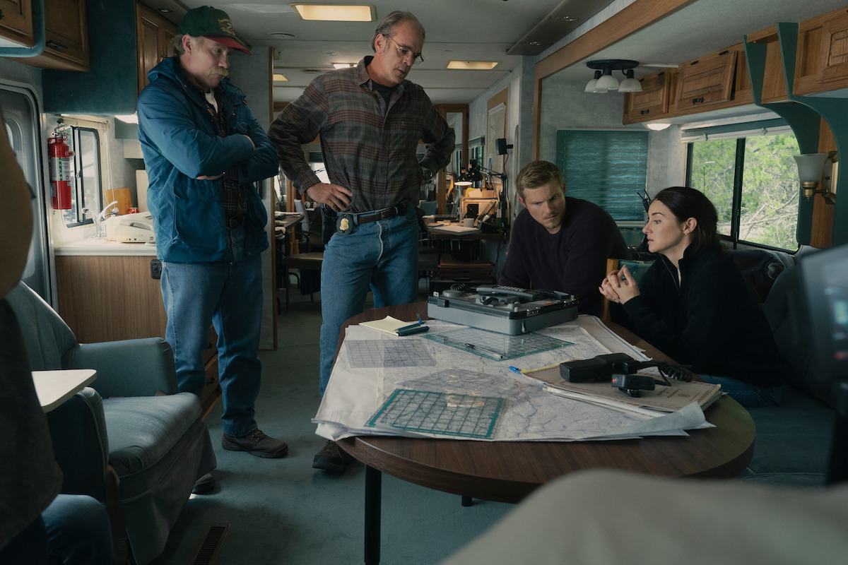 Four people in an RV, gathered around a table covered with maps, papers, and technical equipment, appearing engaged in a serious discussion or investigation.