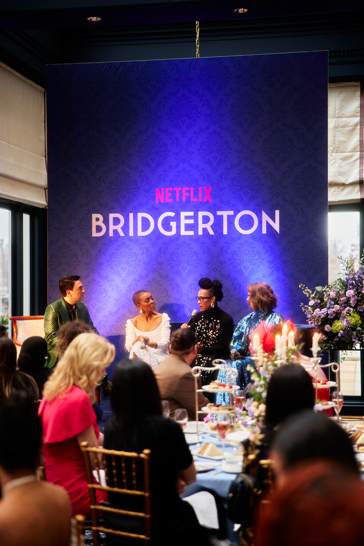 Panelists on stage at a Bridgerton Netflix event, speaking to an audience in a decorated banquet hall with floral arrangements and formal place settings.