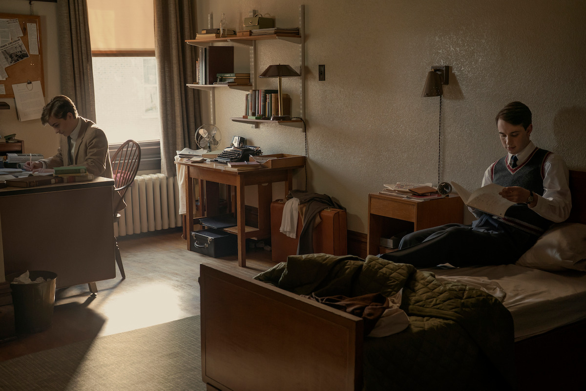 Two young men in a vintage dorm room, one sitting at a desk working, the other sitting on a bed reading a newspaper. The room has a tidy, academic atmosphere with books, lamps, and sunlight filtering through the window.