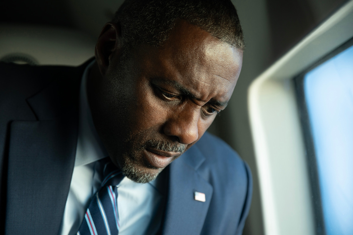 A man in a dark suit and striped tie, looking down thoughtfully inside an airplane, with soft natural light coming through the window, creating a somber and contemplative mood.