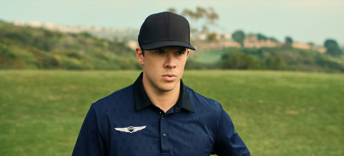 Man in a navy blue collared shirt and black cap standing on a green golf course with trees and distant buildings in the background under a clear sky.