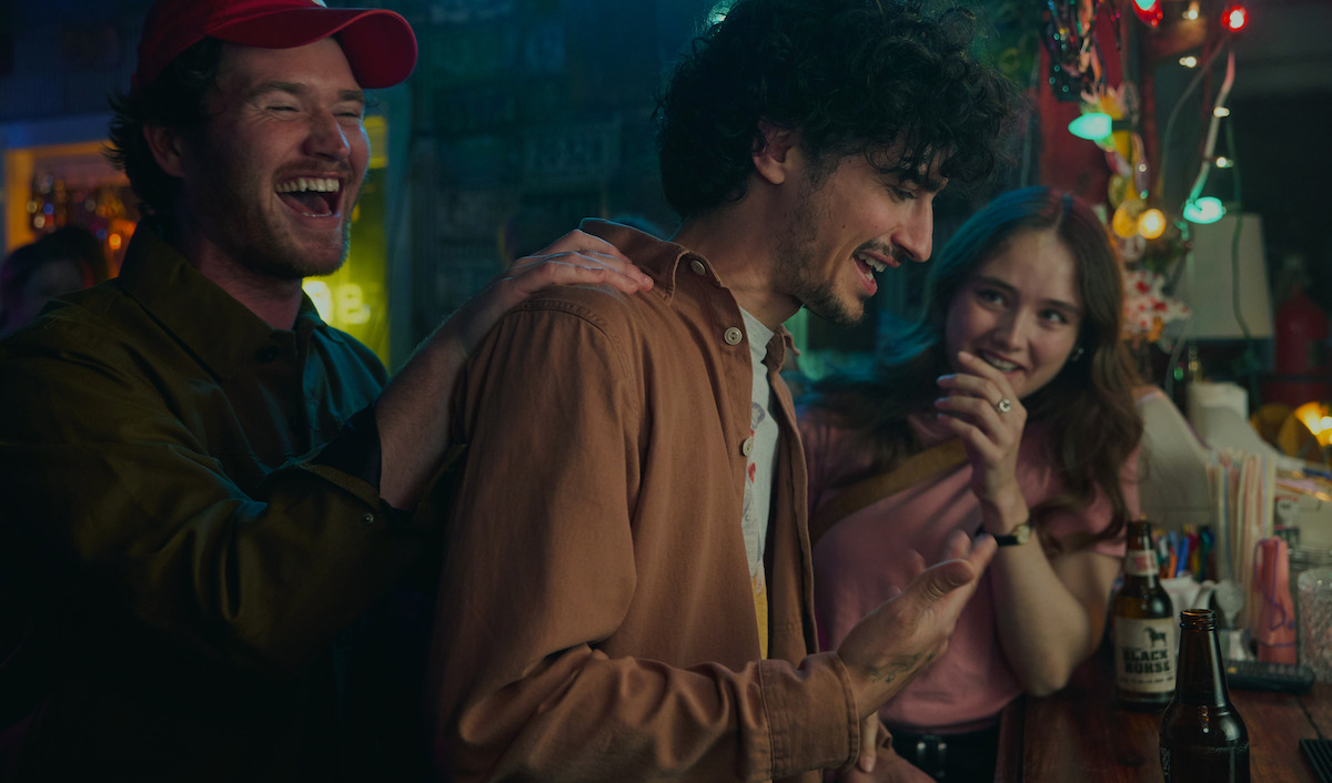Three friends laughing and talking at a bar with drinks, colorful lights, and a casual, lively atmosphere in the background.
