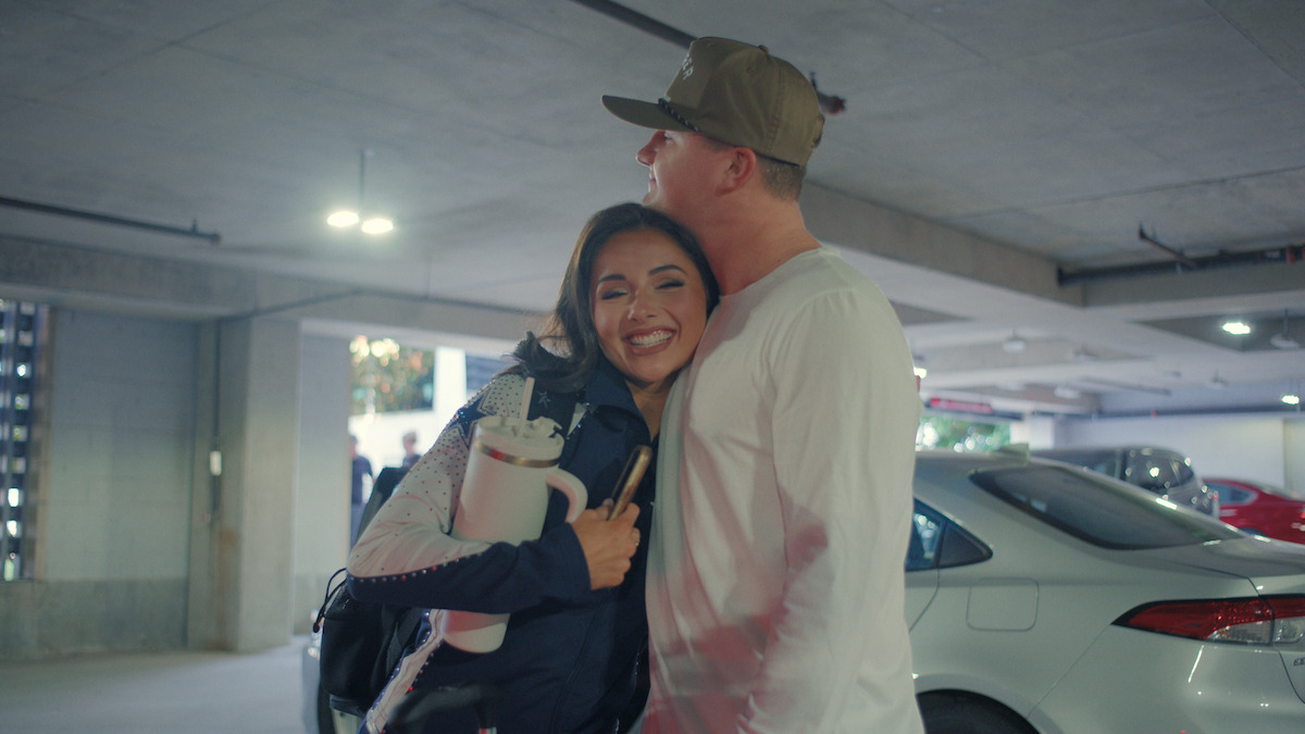 Two people hug and smile in a parking garage, surrounded by parked cars under artificial lighting, suggesting a warm reunion or farewell in an urban setting.