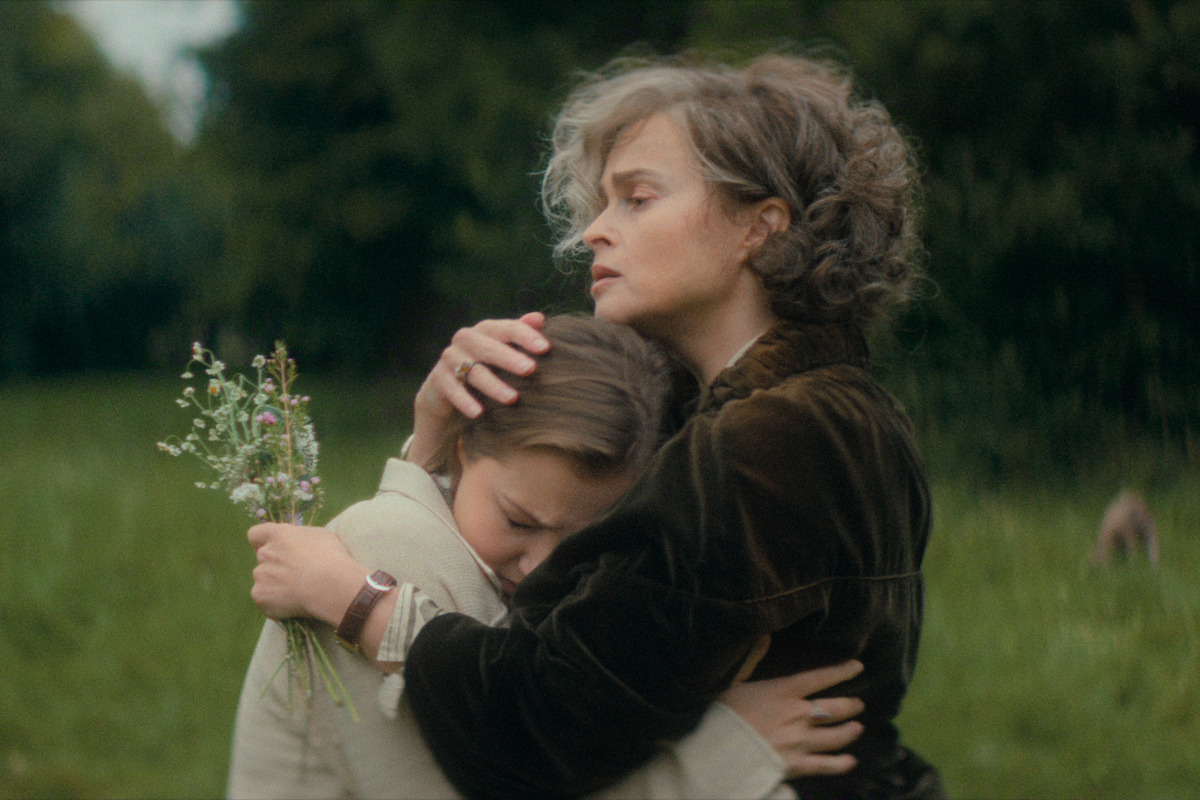 Woman with gray hair in dark coat hugs a sad young girl holding wildflowers in a grassy outdoor field. Both appear emotional, suggesting comfort and support in a natural, green environment.