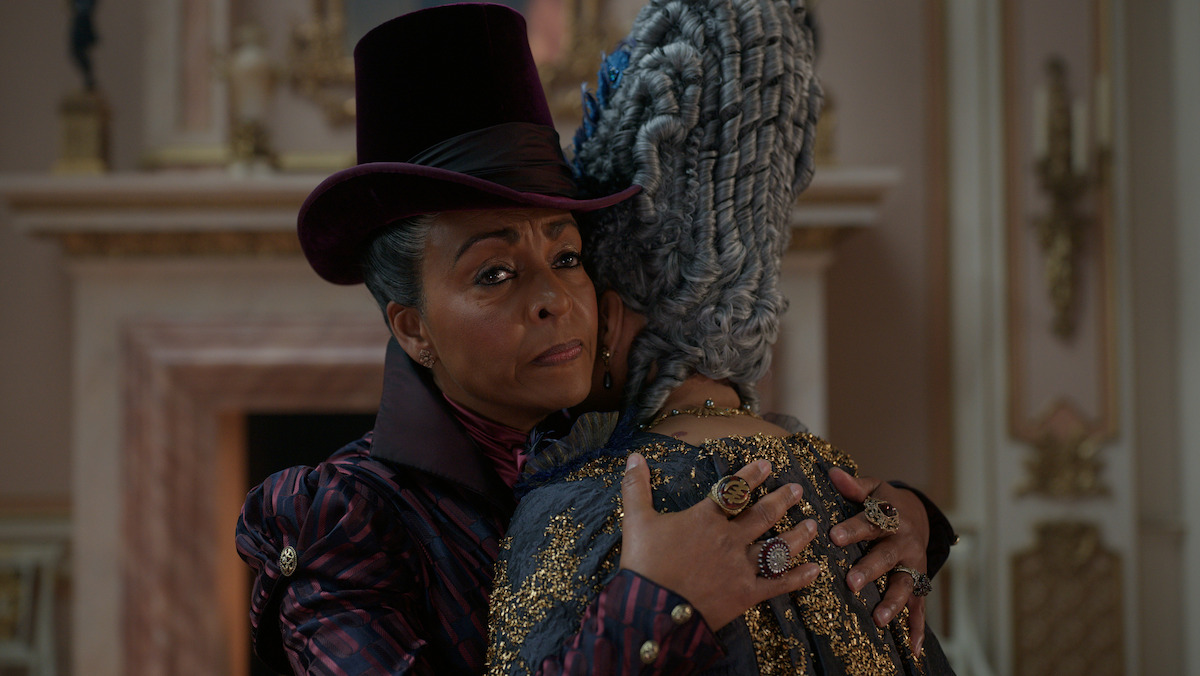 Two elegantly dressed women in period costumes embrace in a lavish room with ornate décor and a fireplace, conveying a moment of comfort or intimacy.