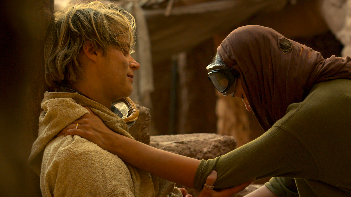 Two people in a desert-like, rustic setting; one sits against a wall wearing a tan cloak, looking tired, while the other, dressed in green with a headscarf and goggles, supports them and appears concerned or comforting.