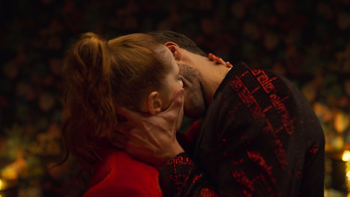 Two people passionately kissing in a dimly lit, intimate setting with floral wallpaper in the background and warm, ambient lighting.