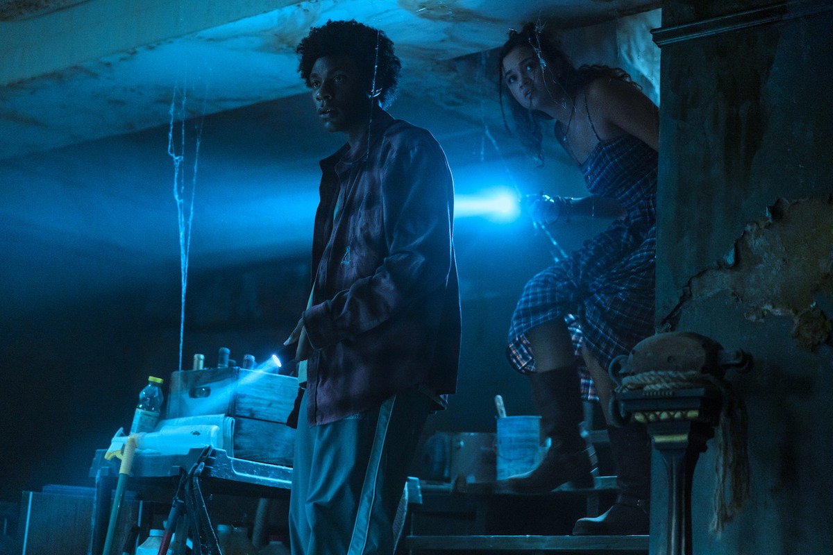 Two people explore a dimly lit, cluttered basement with blue-tinted lighting, holding flashlights and looking cautiously at something off-frame, surrounded by worn concrete and scattered tools.