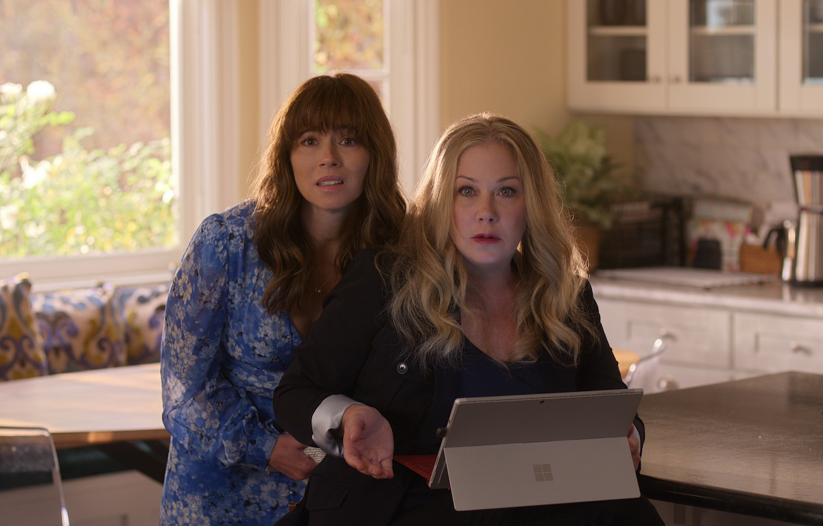 Two women in a bright kitchen, one with long dark hair in a blue floral dress standing, the other with long blonde hair sitting at a counter using a tablet, both looking up with surprised expressions.