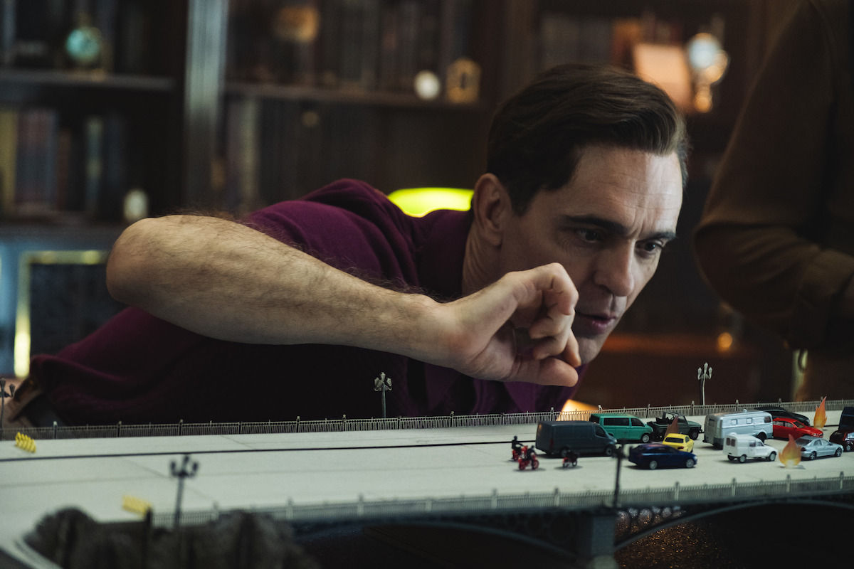 A man in a library or study leans over a detailed model of a bridge with miniature cars and people, carefully arranging the scene with his hand.