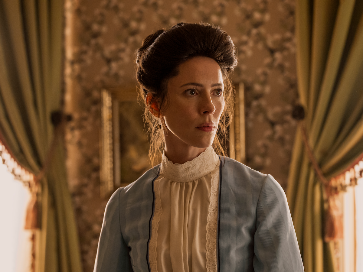 Woman in period clothing with an updo hairstyle stands indoors beside green curtains and patterned wallpaper, with framed artwork in the background. Warm light streams through the windows, creating a vintage, historical atmosphere.