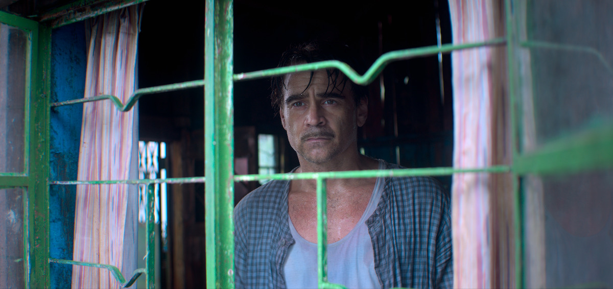 A man with a somber expression stands indoors behind a green metal window, looking outside through rain-streaked glass, with pink curtains framing him, creating a mood of isolation and melancholy.
