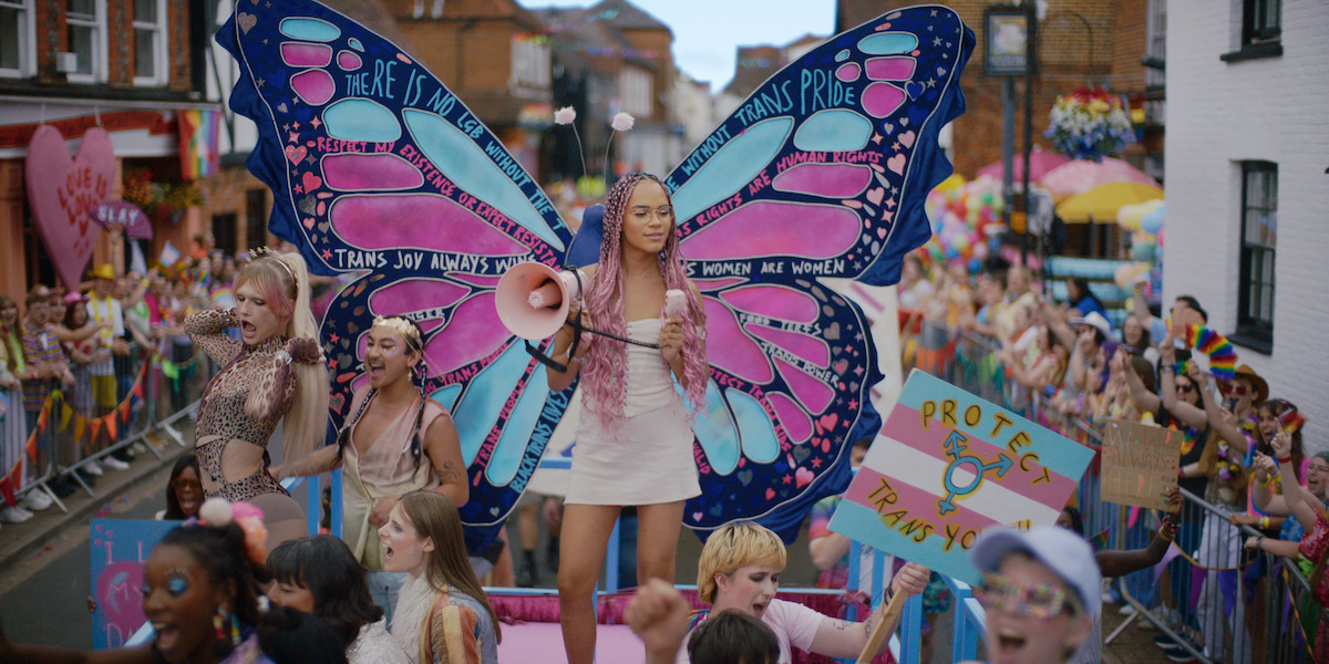 Person with butterfly wings stands on a parade float at a vibrant pride event, surrounded by cheering crowd, colorful signs supporting trans rights, and festive decorations on a city street.