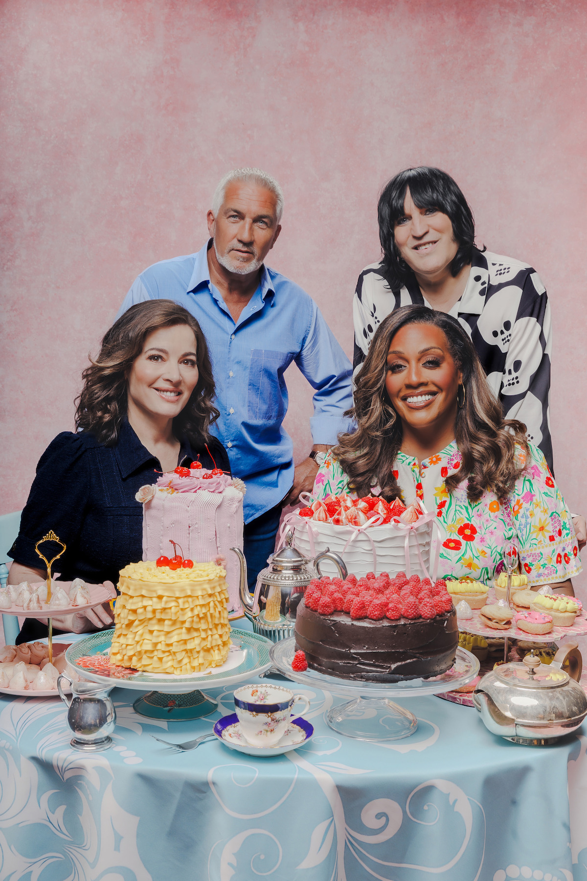 Four people pose behind a table decorated with a blue tablecloth, assorted cakes, pastries, and tea cups, set in a colorful, festive indoor environment with a pink backdrop.
