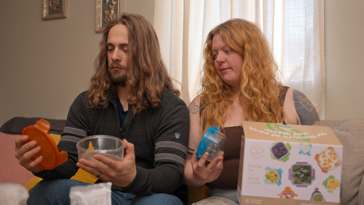 Two people sitting indoors on a couch, examining plastic containers and lids. The man holds an orange lid and clear bowl, the woman holds a blue lid and container. A product box with images is beside them. Light curtains and framed art on wall.
