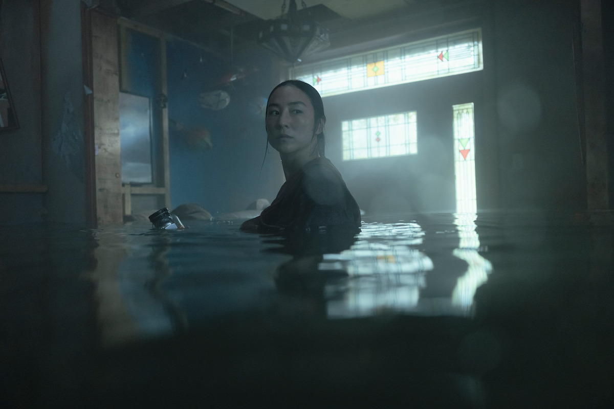 Greta Lee as Anne standing in murky water inside a flooded room with stained glass windows, gloomy lighting, and partially submerged objects creating an eerie, tense atmosphere.