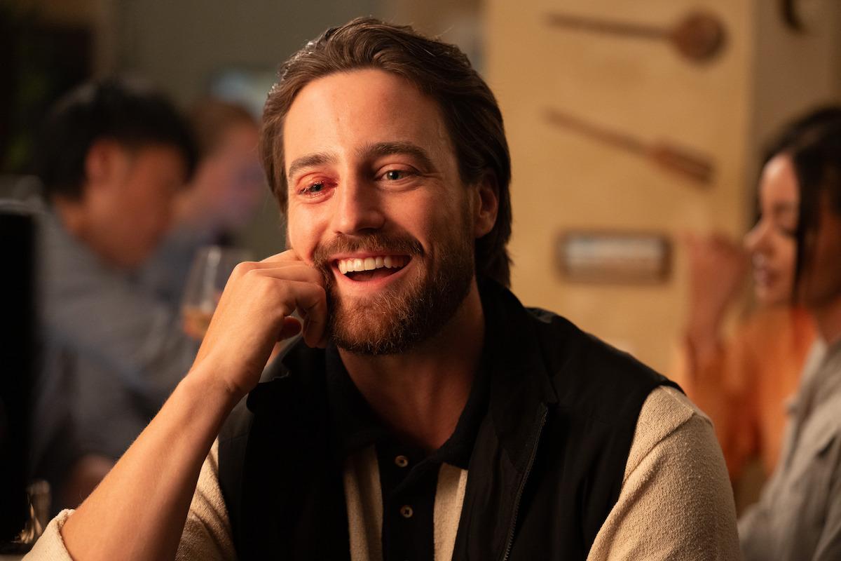 A smiling man with a reddish mark near his eye sits at a cozy, warmly lit restaurant or bar, engaging in conversation with people in the blurred background.